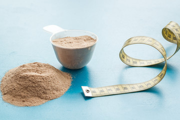Whey protein powder in plastic scoop near measuring tape on blue background