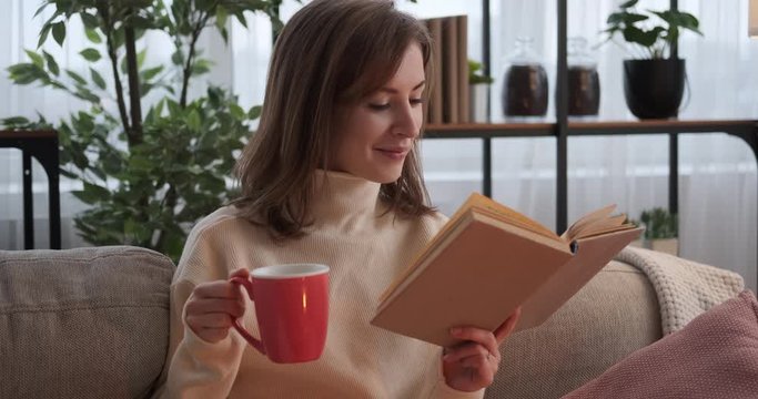 Happy woman reading a book and drinking coffee on sofa at home
