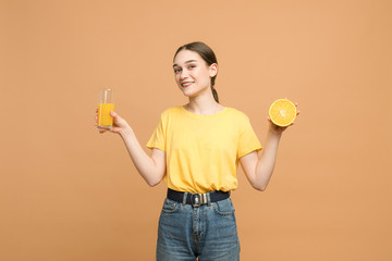 Happy attractive brunette girl in yellow shirt holds sliced orange in right hand and glass of juice in left, smiling and looking at the camera