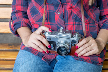 Girl in blue jeans and checkered shirt is sitting on the wooden bench and holding an old film camera in her hands.