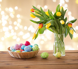 easter, holidays and object concept - colored eggs in wicker basket and tulip flowers in vase on wooden table over festive lights on beige background