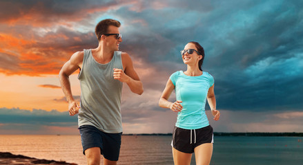 fitness, sport and healthy lifestyle concept - happy couple in sports clothes and sunglasses running over sea and sunset sky background