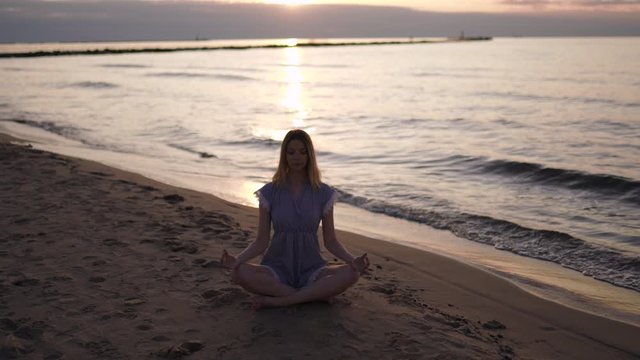 Caucasian blonde woman meditates at sandy beach wearing blue summer dress during sunrise. Wide panning right shot