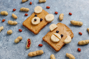 Funny bear and monkey face sandwich with peanut butter, banana and black currant,peanuts on grey concrete background,top view