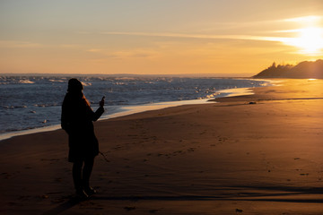 Silhouette of woman with smartphone on the beach with unfocused sea, sun and sky on evening background.