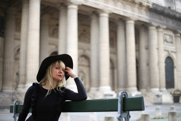 Beautiful long-legged blonde in a short black dress and hat sits on a bench in Paris