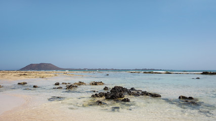 Fototapeta na wymiar Hot summer views towards the small island of Los Lobos few miles north from Corralejo resort, with calm waves, clear and shallow waters and tropical atmosphere, in Fuerteventura, Canary Islands, Spain