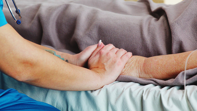 Close up shot of young caucasian assitant taking the hand of an old lady lying in hospital bed