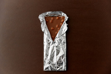 sweets, confectionery and food concept - milk chocolate bar with nuts in foil wrapper on brown background