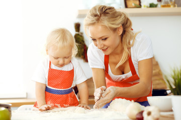 Little girl and her blonde mom in red aprons playing and laughing while kneading the dough in kitchen. Homemade pastry for bread, pizza or bake cookies. Family fun and cooking concept