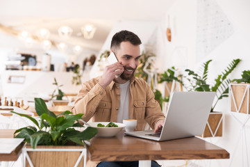 Young handsome bearded man working in his laptop talking on smartphone in his hand during lunch...