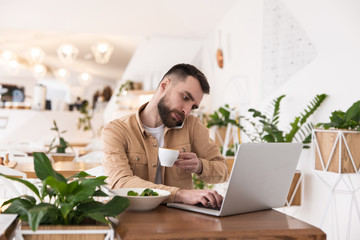 Young handsome bearded smiling man looks busy typing in laptop talking on phone during lunch break...
