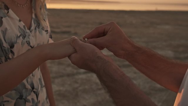 A cropped view of hands when the man is putting on a ring to a woman while making a proposition of marriage on the sunny beach near the lake