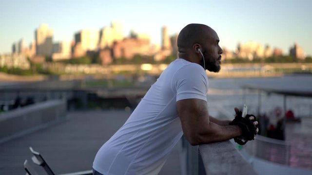 Young dark  skinned male jogger with muscular body shape in active wear resing on bridge and listening music via earphones and smartphone