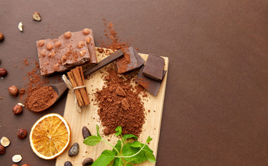 sweets, confectionery and culinary concept - chocolate with hazelnuts, cocoa beans, and powder with dry orange and cinnamon on wooden board with spoon on brown background