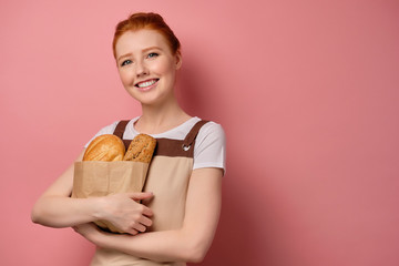 A red-haired girl with collected hair stands on a pink background in an apron and smiles, clutching a paper bag with bread. 