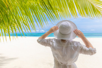 Fototapeta na wymiar Woman in white beach dress and beach hat under palm leaves. Amazing summer vacation and travel background. Woman on beach, summer holiday view
