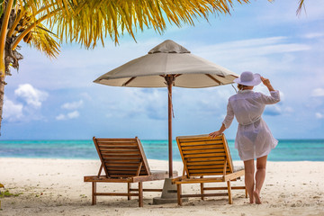 Woman at beautiful beach in white dress and white hat under palm trees relaxing on lounge chair. Tropical paradise island, summer vacation or holiday. Blue sea, white sand, happy summer mood