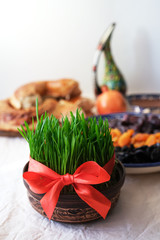 Young green wheat with red ribbon, traditional decoration of holiday Navruz. Background - national dessert of Central Asia called Sumalak, lavash bread, assortment of dry fruits, pomegranate.