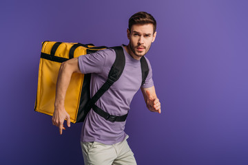 concentrated delivery man running in a hurry while carrying thermo backpack isolated on purple