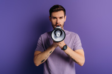 handsome young man speaking in megaphone while looking at camera on purple background