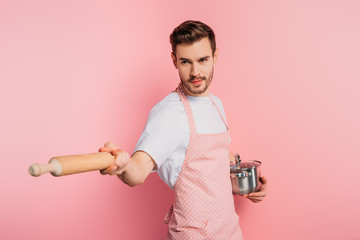 concentrated young man in apron imitating fencing with saucepan and rolling pin on pink background