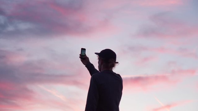 Silhouette of young hipster man make photo or video of epic pink and purple sunset. Social media influencer or blogger. Amazing cinematic view of nature sky. Romantic wanderlust concept