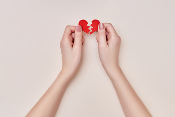 Female hands holding two parts of a broken red paper heart on the biege background