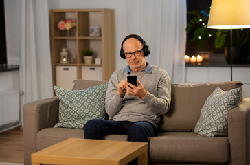 Obraz premium technology, old people and lifestyle concept - happy senior man with smartphone and headphones listening to music at home in evening