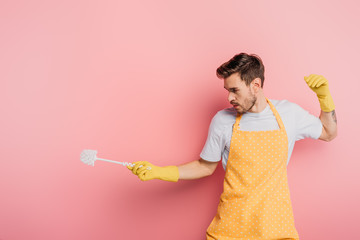 young man in apron and rubber gloves imitating fencing with plunger on pink background