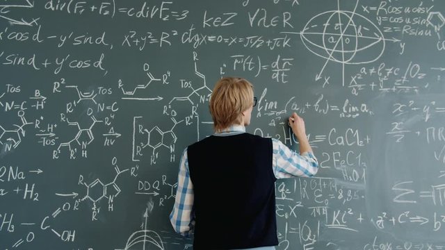 Back view of young man maths teacher writing equations on chalkboard in classroom busy with calculations. Occupation, lifestyle and people concept.