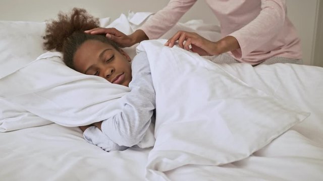 Pleased african woman covering her little sleeping pretty daughter with blanket in bed at home