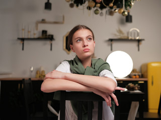 Young girl in a white T-shirt and a green cardigan draped over her shoulders sits on a wooden chair...
