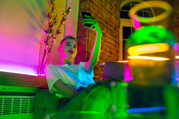 Selfie. Cinematic portrait of stylish woman in neon lighted interior. Toned like cinema effects, bright neoned colors. Caucasian model using smartphone in colorful lights indoors. Youth culture.