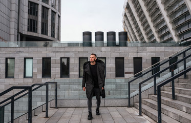  A guy, a model with brown hair in a black coat, black jeans and sneakers, poses against the backdrop of architecture. Man portrait