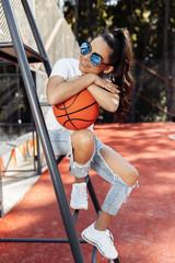 Brunette young woman dressed in casual modern clothes posing with basket ball on referee chair at...