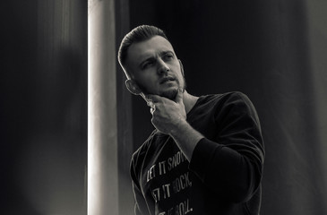 A guy, a model with brown hair in a black coat, black jeans and sneakers, posing against a background of architecture. Black and white man portrait