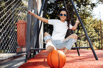 Adorable young woman dressed in casual modern clothes posing with basketball at the stadium.
