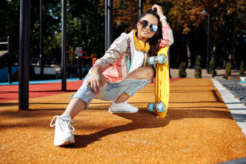 Modern brunette young woman sitting and listening music holding yellow skateboard or pennyboard at...