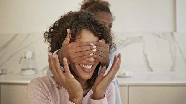 Pleased little girl covering eyes to her mother behind which sitting by the table on kitchen at home