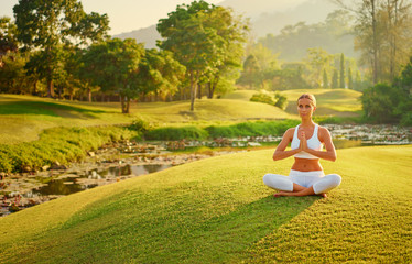 Obraz na płótnie Canvas Yoga at park with view of the mountains, with sunlight. Young woman in lotus pose sitting on green grass. Concept of calm and meditation.