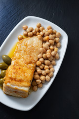 fried cod fish with chick pea and olives on white dish on ceramic background