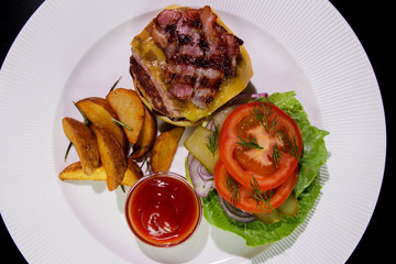Burger with fried bacon, french fries, vegetables and ketchup on a white plate.View from above.Close-up.Unhealthy food.