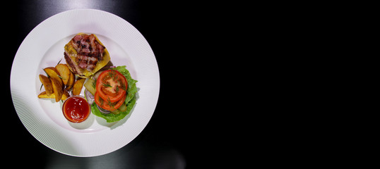 Burger with fried bacon, french fries, vegetables and ketchup on a white plate.Copy space.Isolated on a black background.View from above.Horizontal composition.Unhealthy food.