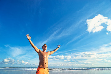 Fototapeta na wymiar Victory and freedom. Young handsome strong man raising hands up on the beach against sky.