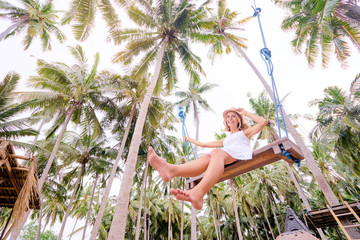 Vacation concept. Happy young woman in white dress and hat swinging at palm grove.