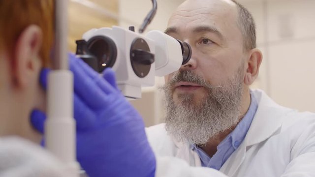 Professional senior optometrist in disposable gloves and lab coat looking through oculars of slit lamp and telling something while examining eyes of little boy