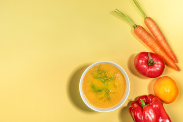Fresh vegetable cream soup and ingredients: carrots, tomatos and sweet pepper for its preparation on yellow background.