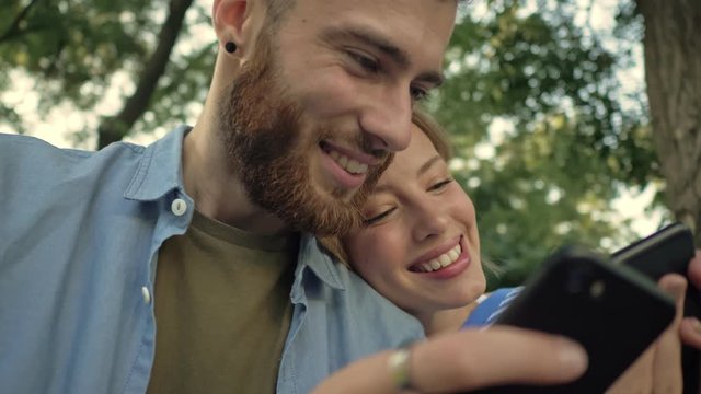 Close-up view of a young smiling couple is using smartphones while sitting on the bench with electric kick scooters at a park landscape