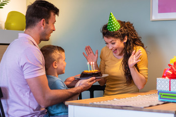 Smiling family celebrating a birthday together at home, father and son surprise mother with cake.